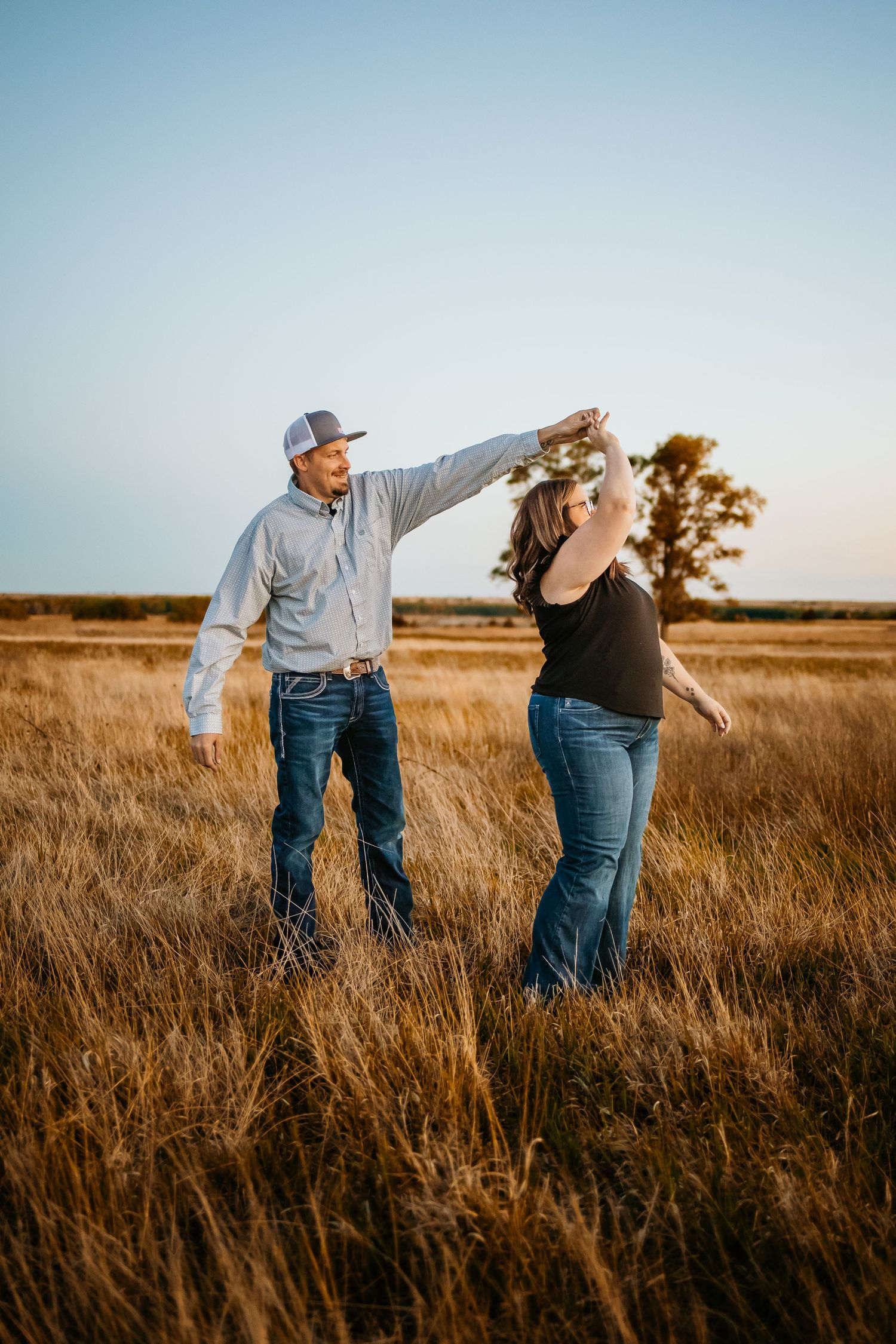 Couple dancing in golden prairie field at sunset, man in cap twirling woman in jeans under his raised arm.