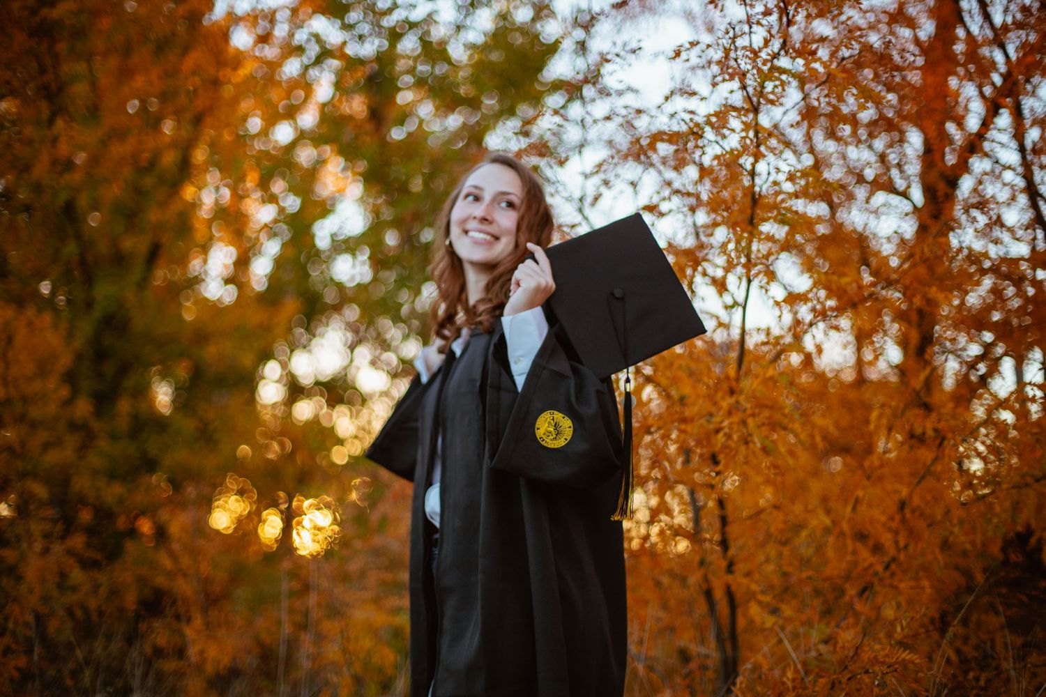 Graduate in black cap and gown holds diploma while standing against bright autumn trees at sunset.