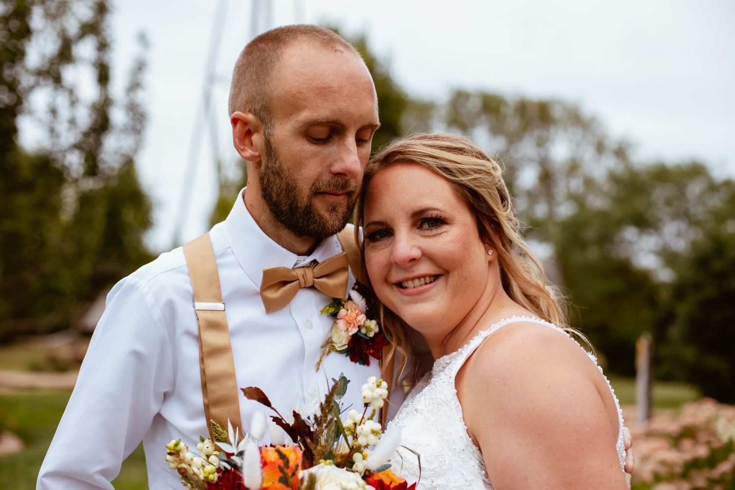 A couple in wedding attire poses with autumn-themed floral bouquet outdoors.