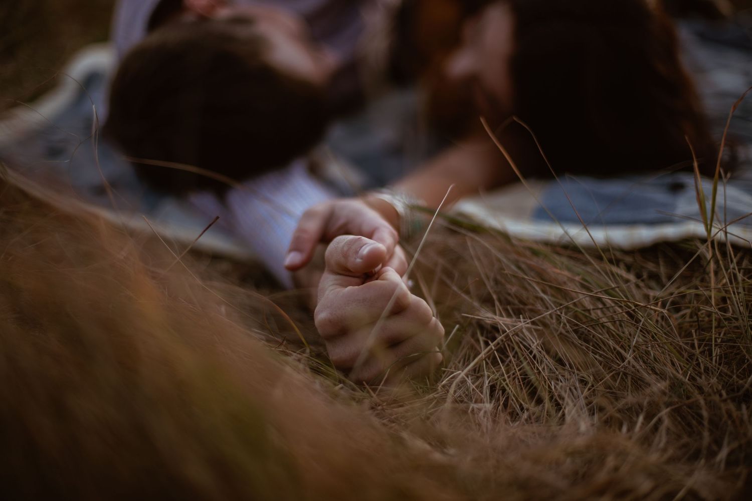 A close-up photo of clasped hands resting in dry autumn grass during sunset.