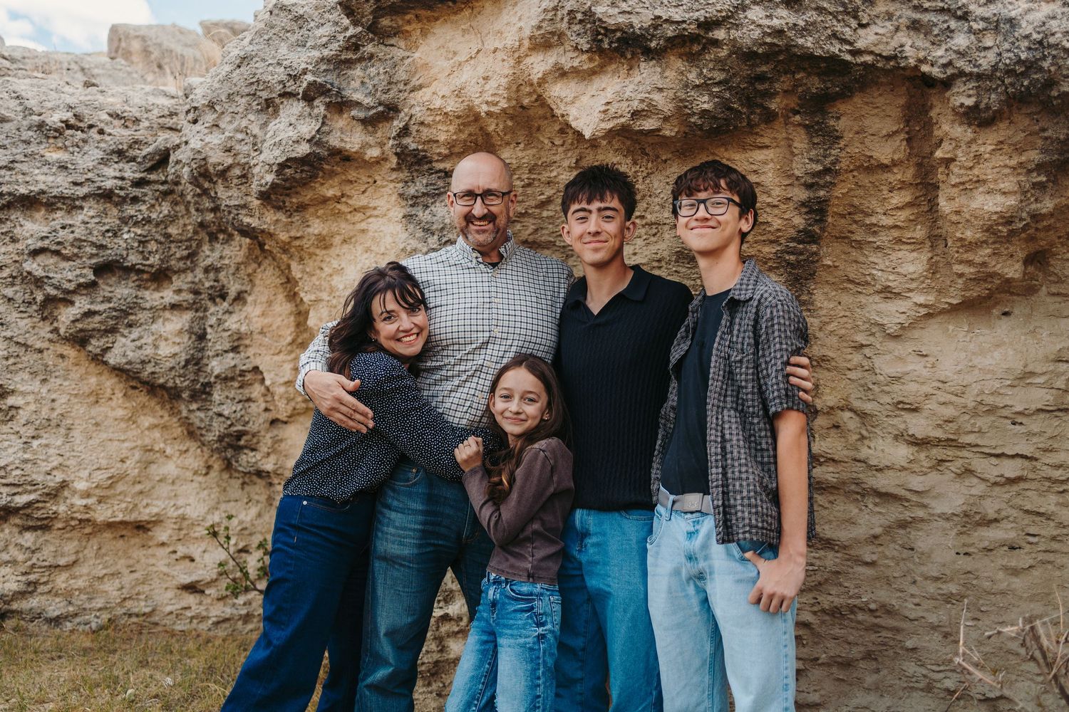 Family of five smiling together outdoors near rocky cliff, casual portrait with children and adult man.