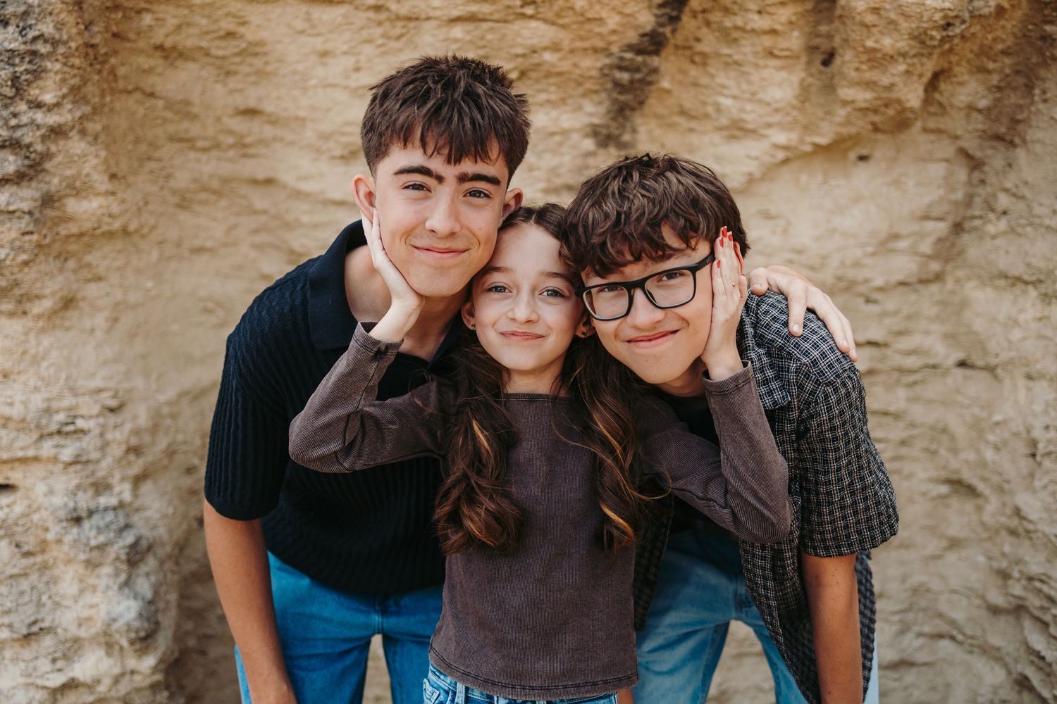 Three smiling siblings pose together against a sandy rock wall, two older boys flanking a young girl in a warm outdoor portrait.