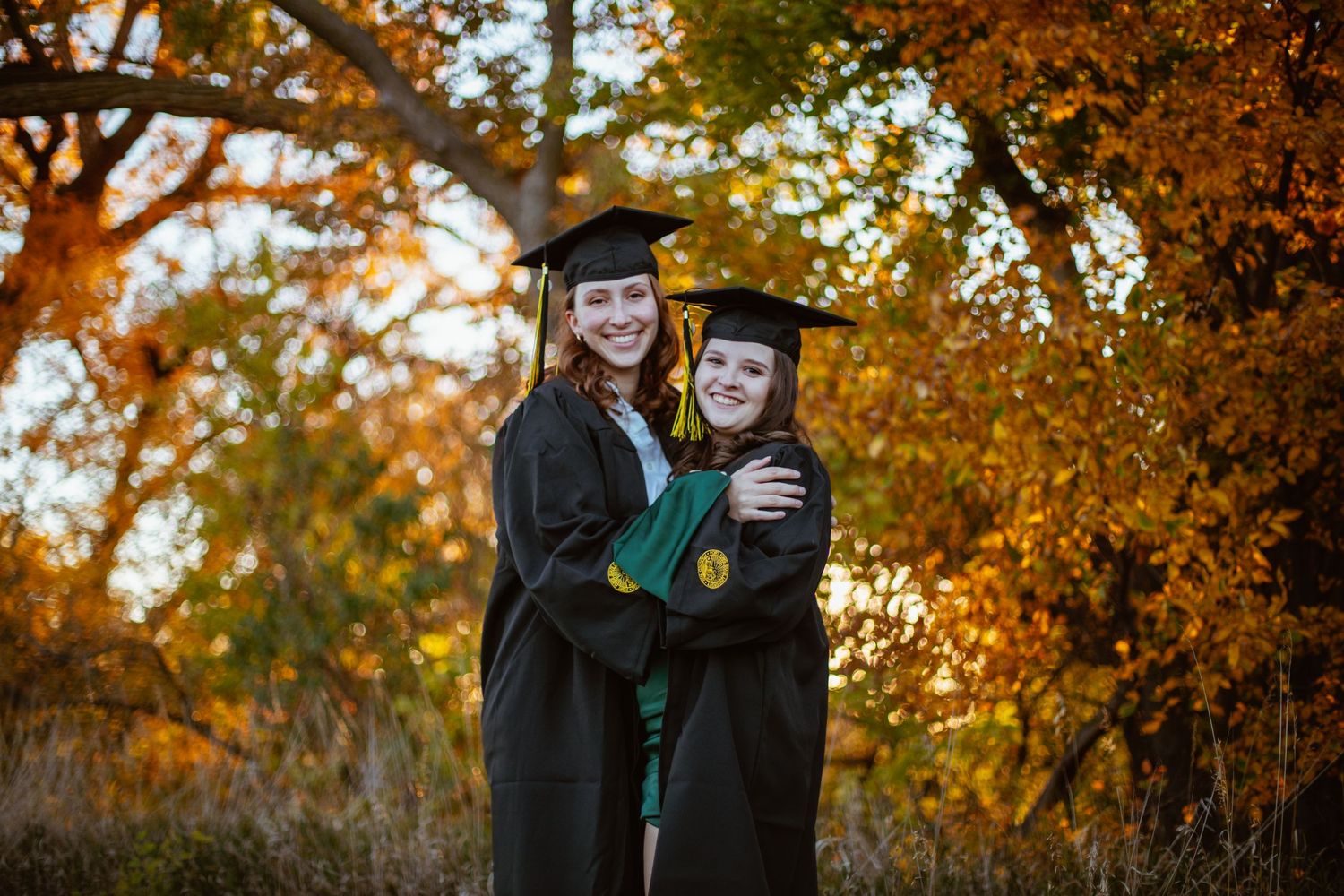 Two graduates in black caps and gowns embrace under golden autumn trees.