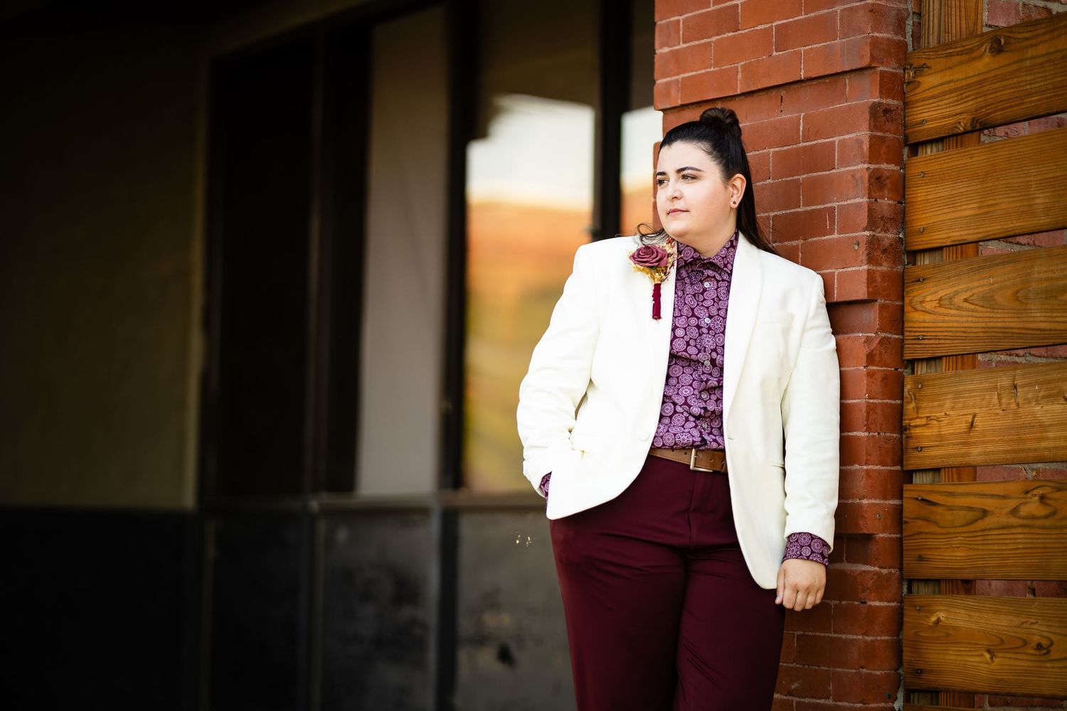 A person in a white blazer and burgundy pants stands against a brick and wood wall in formal attire.
