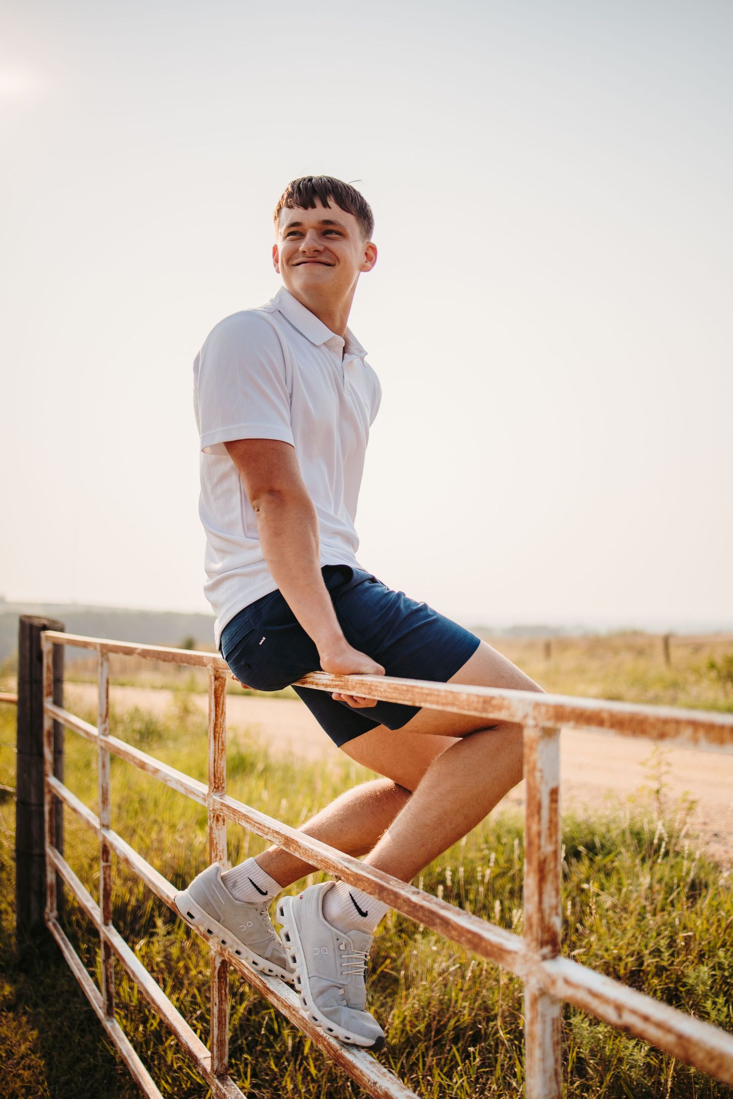 Person in white polo and blue shorts sits casually on rustic wooden fence during golden hour.