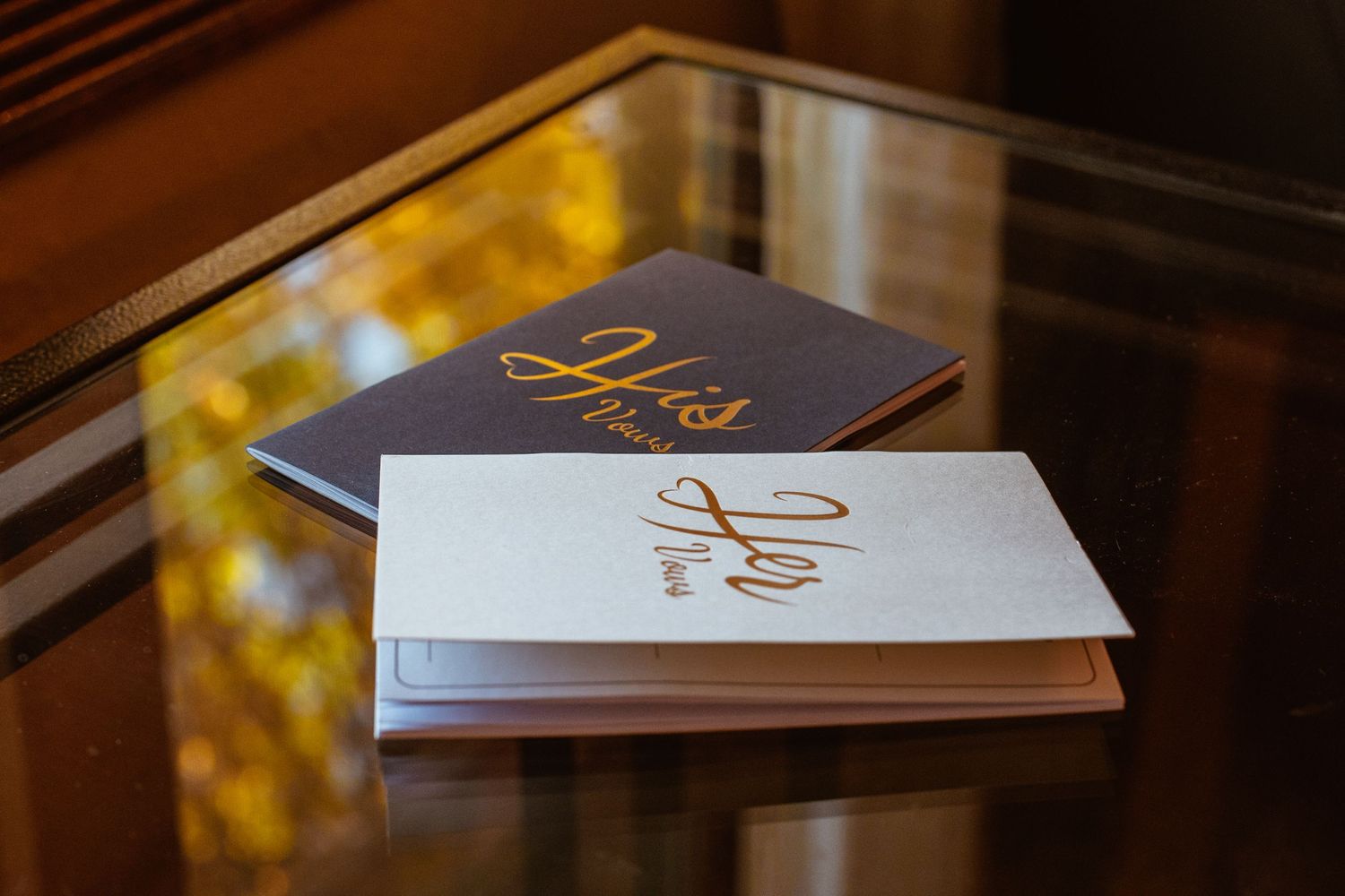 Gold foil wedding vows booklets stacked on reflective surface with warm lighting.