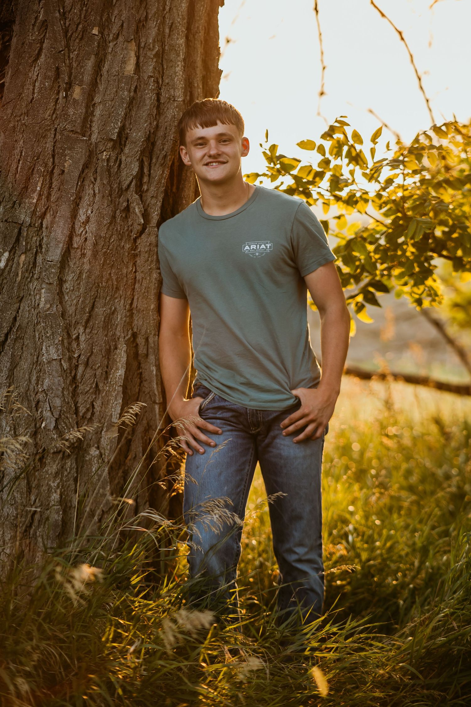 Young man in Ariat t-shirt and jeans leaning against a tree in golden hour sunlight outdoors.