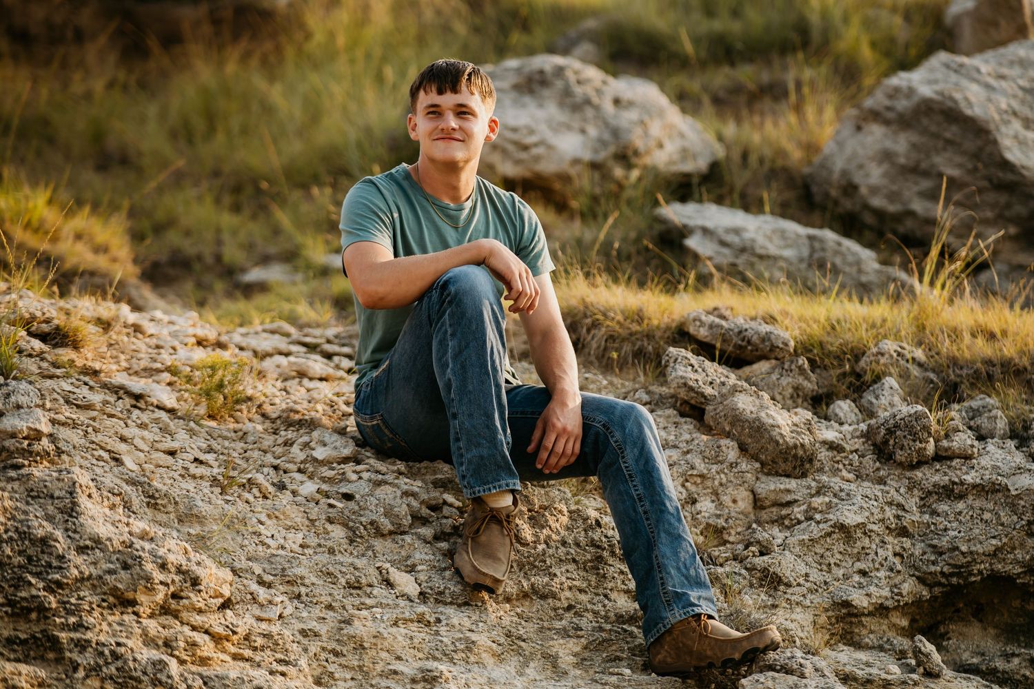A person in casual clothing sits on rocky terrain during golden hour in a natural outdoor setting.