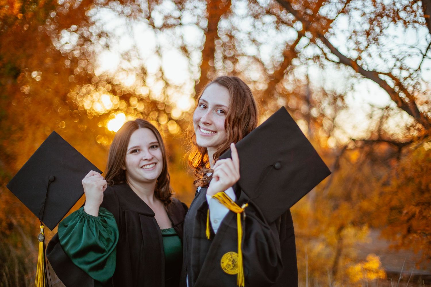 Two graduates in black and green regalia hold diplomas while celebrating under fall foliage.