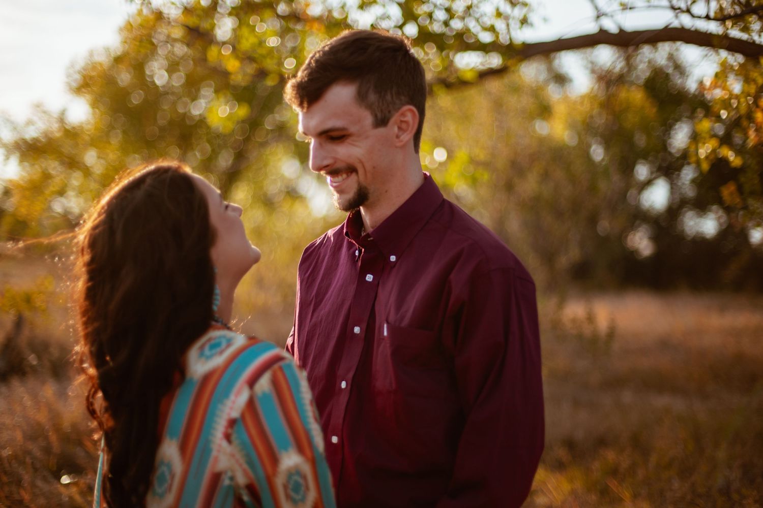 A couple laughs together in a field during golden hour wearing burgundy and blue.