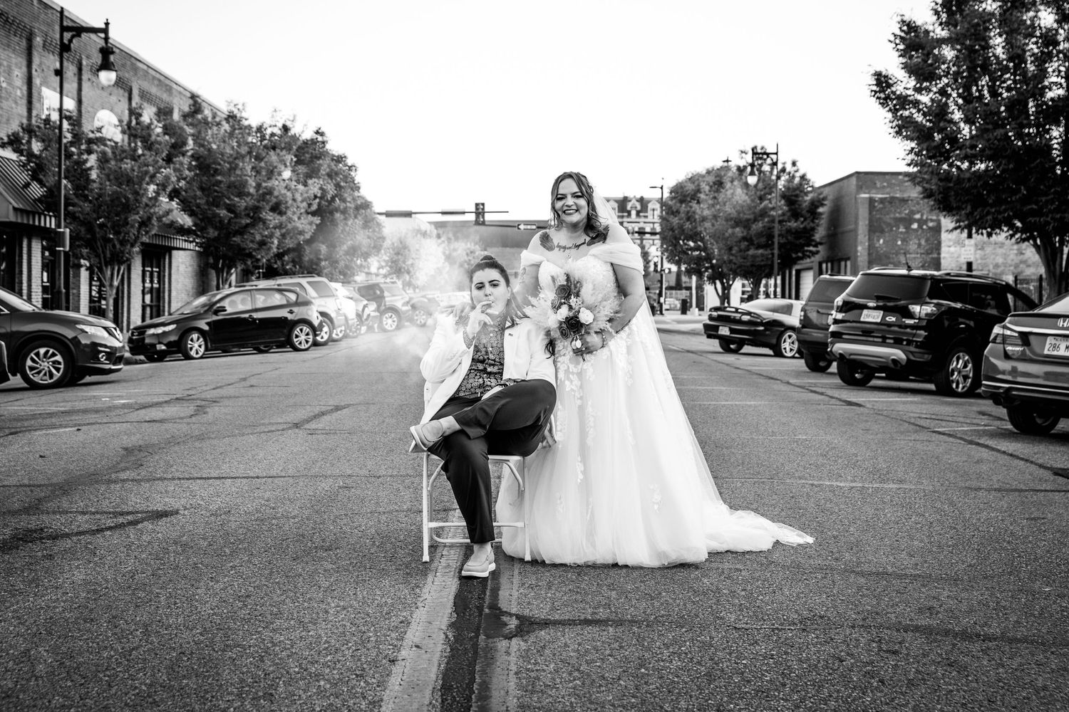 A wedding couple poses on a street lined with parked cars in black and white photography.