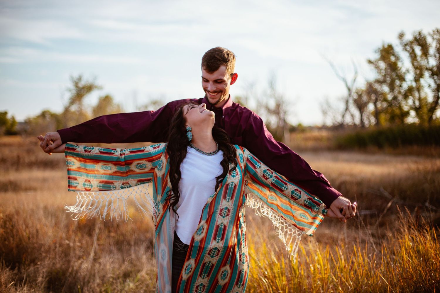 Couple wrapped in patterned blanket stand together in autumn field.
