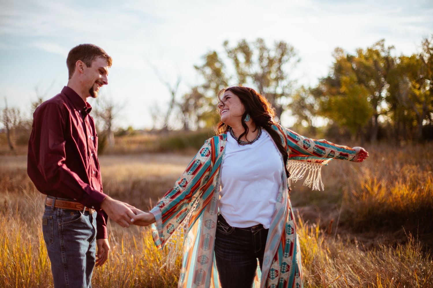 A couple laughs together in a golden field during sunset while holding hands.