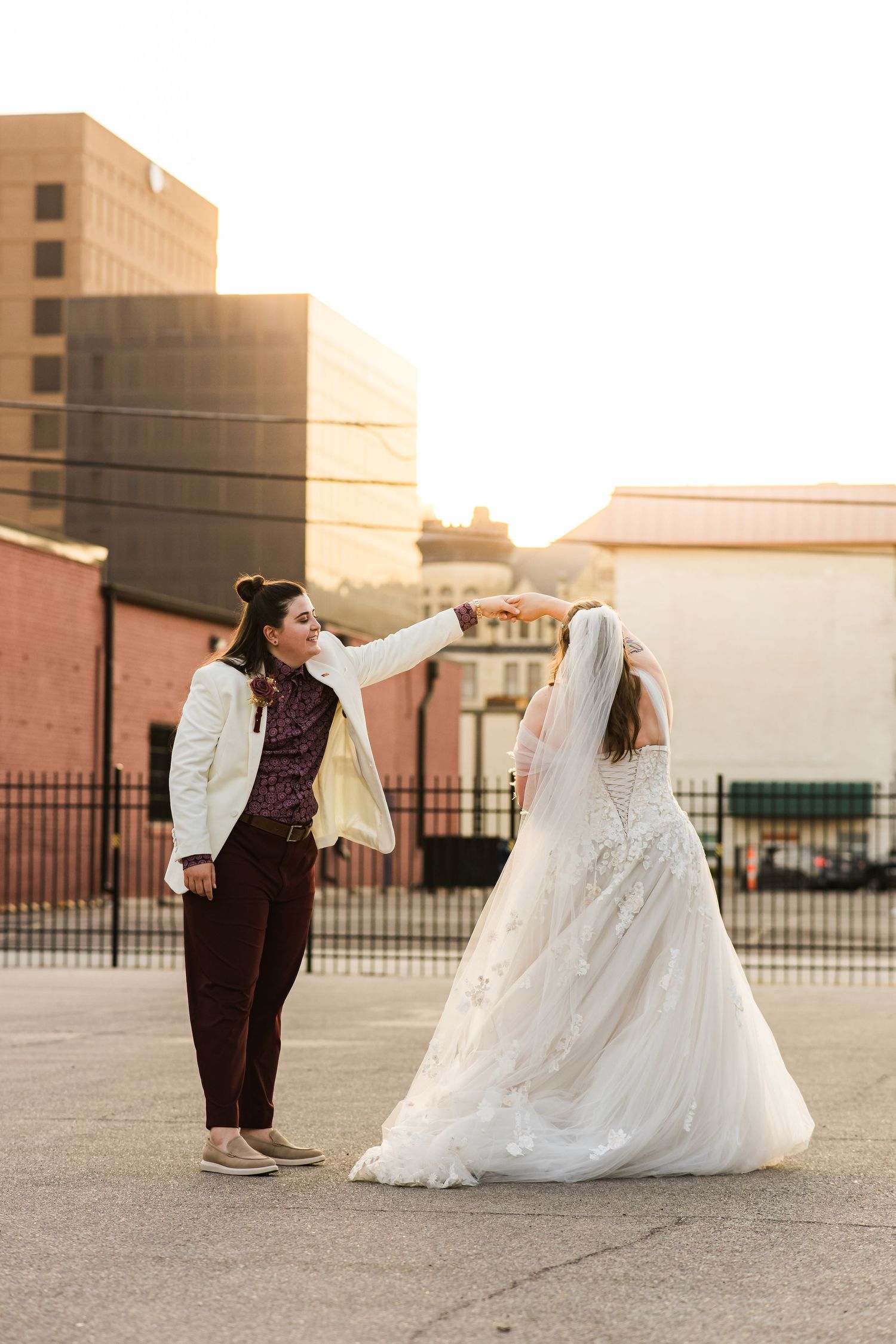A couple in wedding attire poses playfully in an urban setting at sunset.