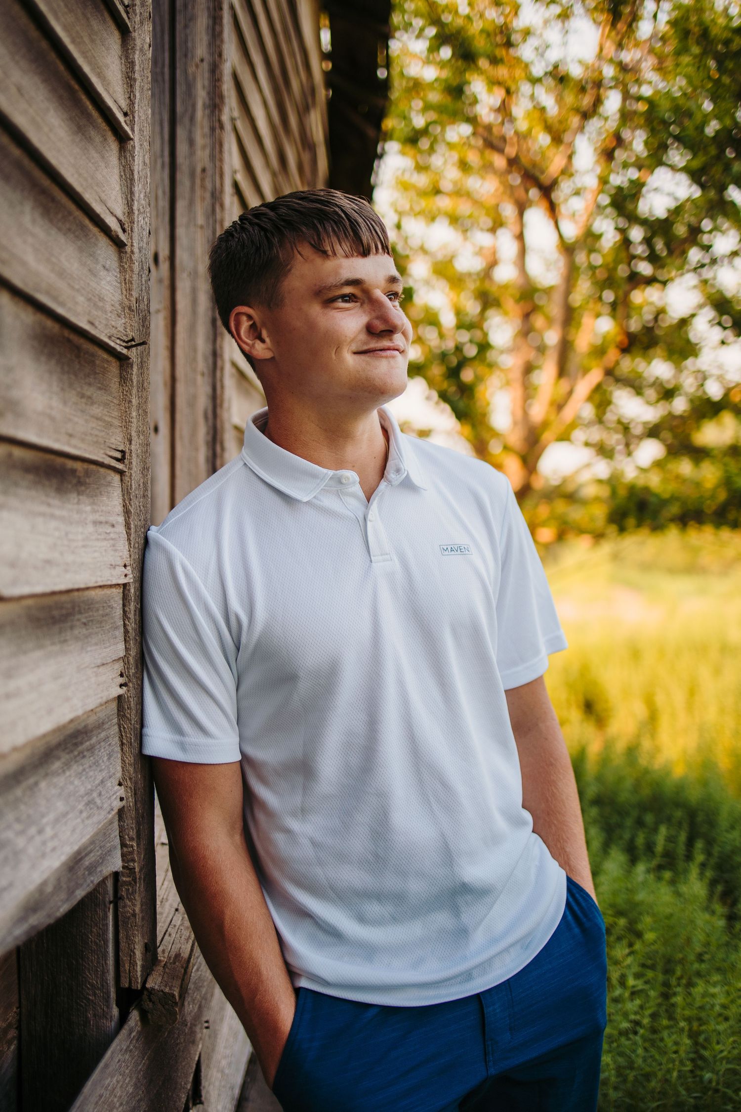 Person in white polo shirt stands against rustic wooden wall in warm evening light.
