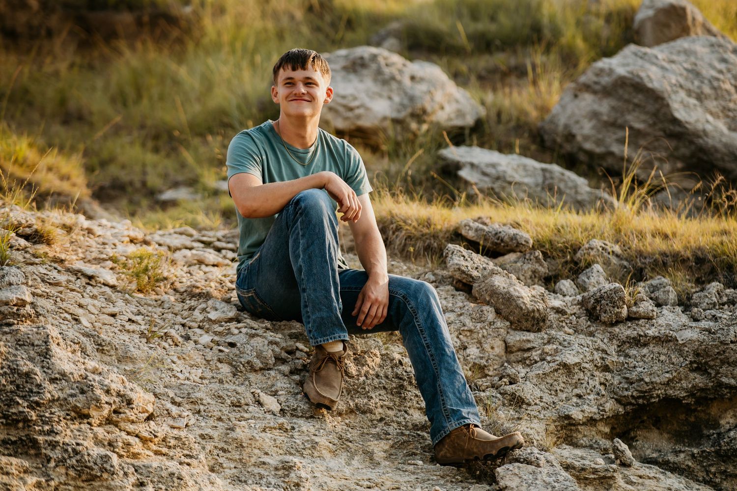 Young man in jeans and green t-shirt sitting on rocky terrain outdoors, smiling in natural golden light.