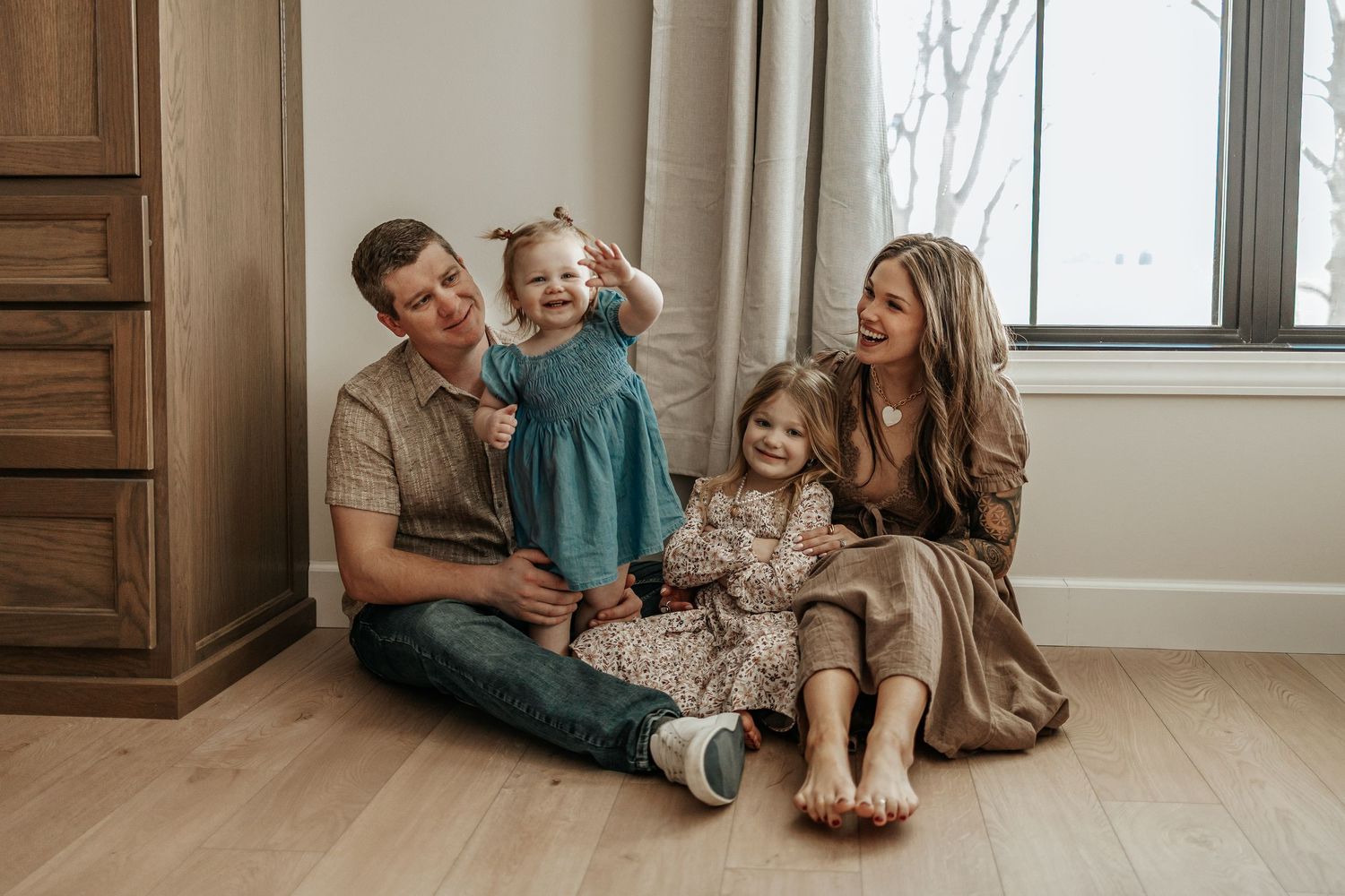Happy family of four sitting on hardwood floor near window, parents smiling with two young daughters in cozy home setting.
