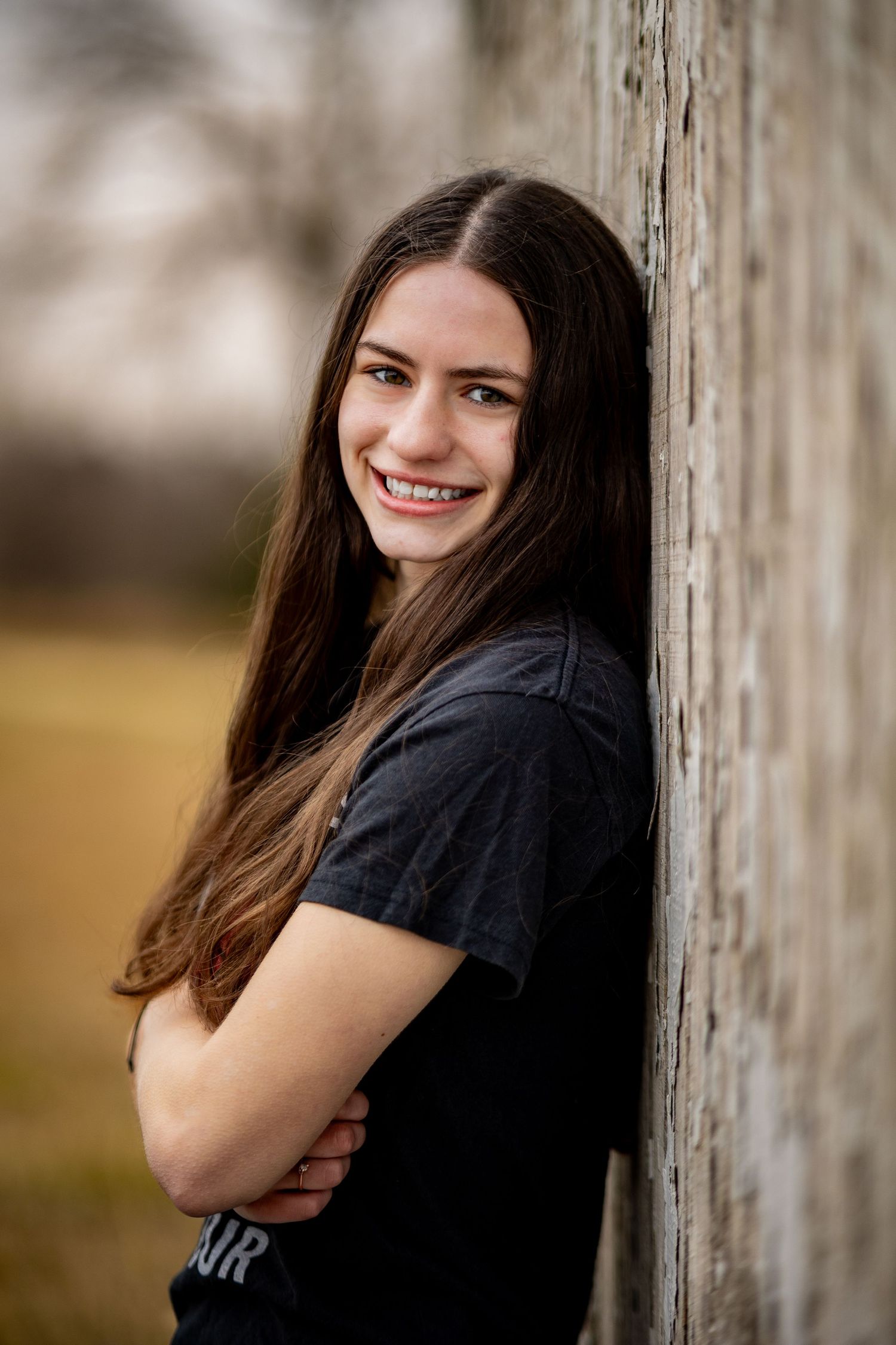 Portrait of someone with long dark hair smiling while leaning against rustic wooden wall.