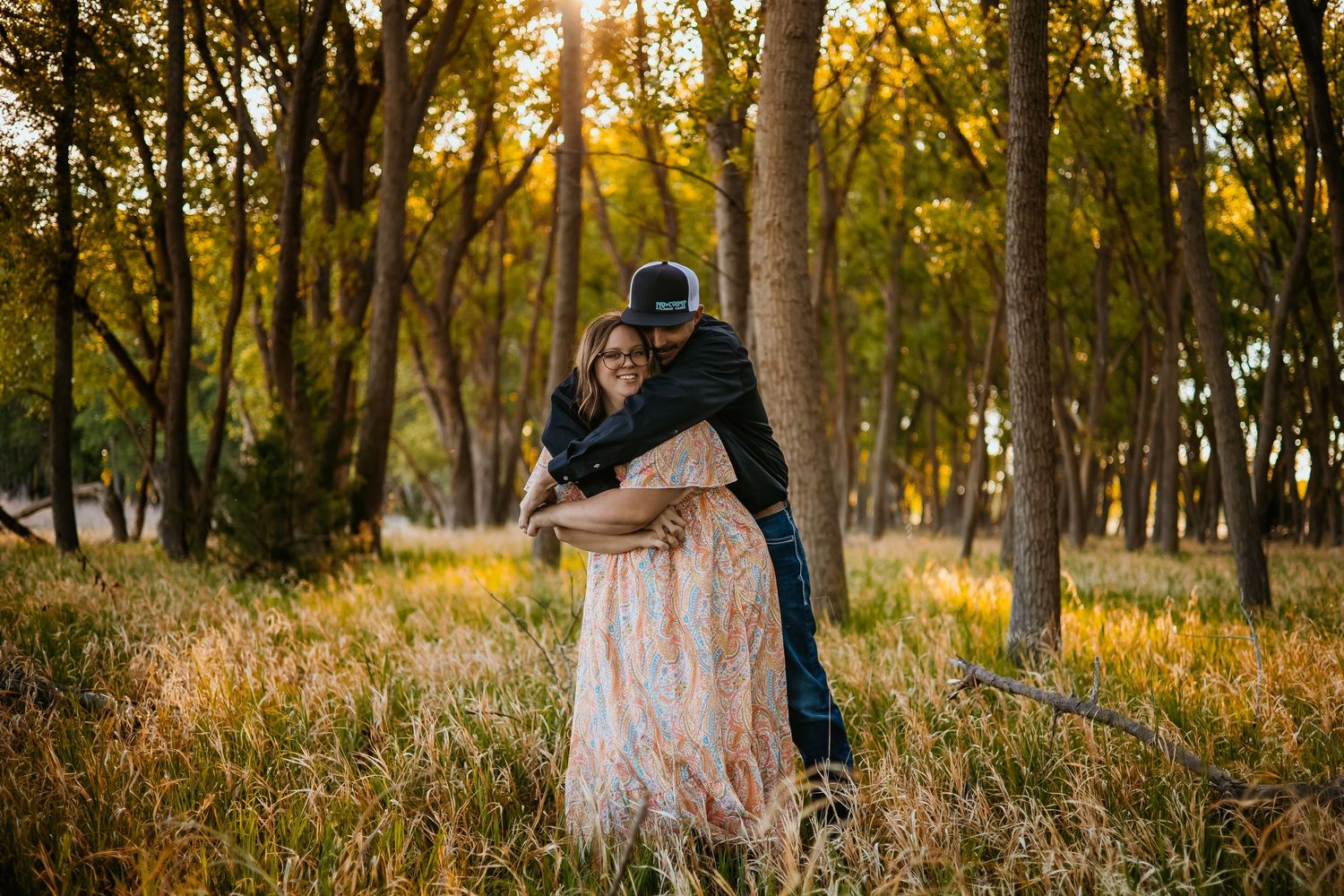 Couple embracing in golden sunlit forest meadow, woman in floral dress, man in black hat, romantic outdoor engagement photo.