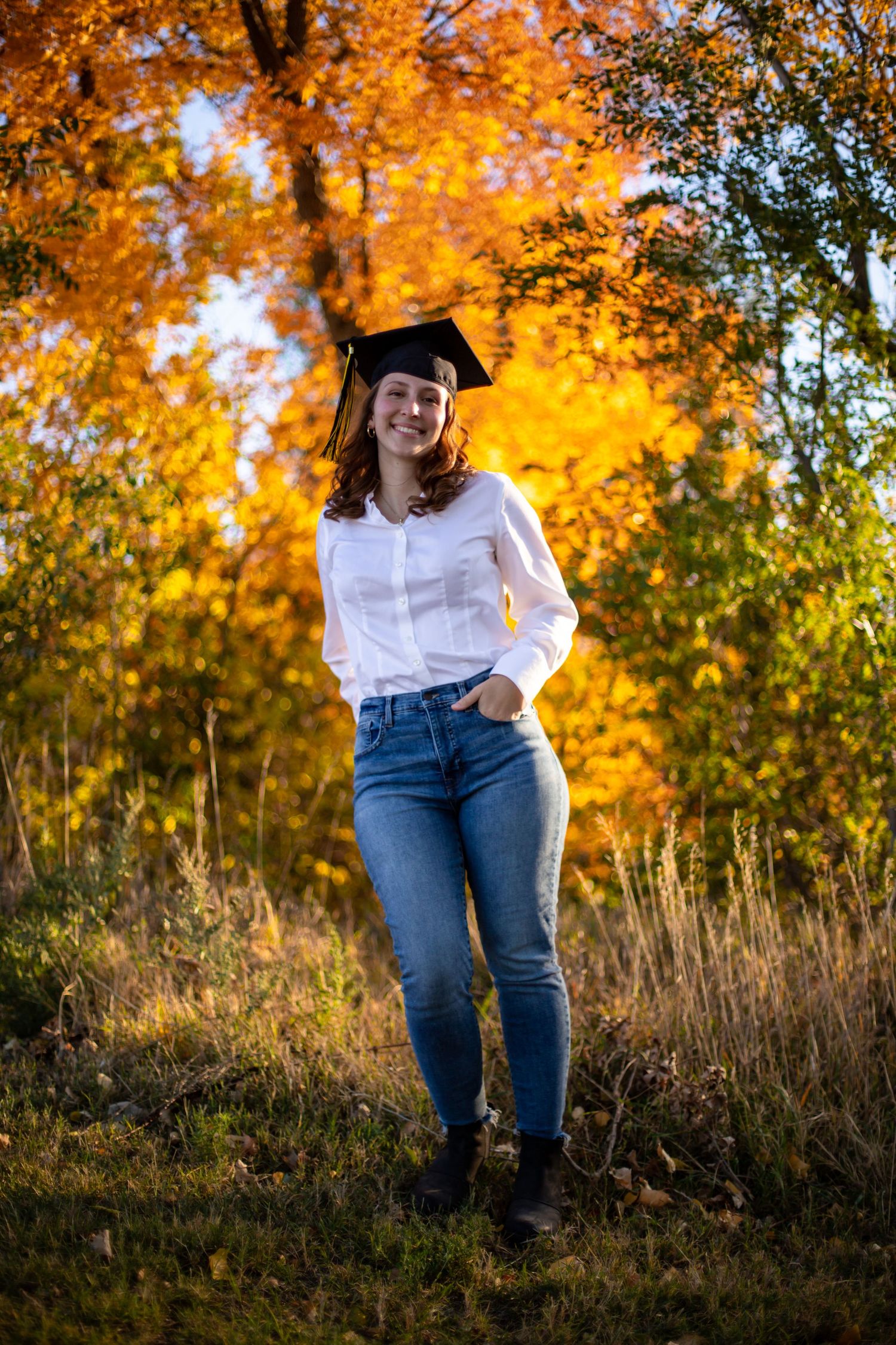 Graduate in white shirt and jeans poses amidst vibrant fall foliage with a graduation cap.
