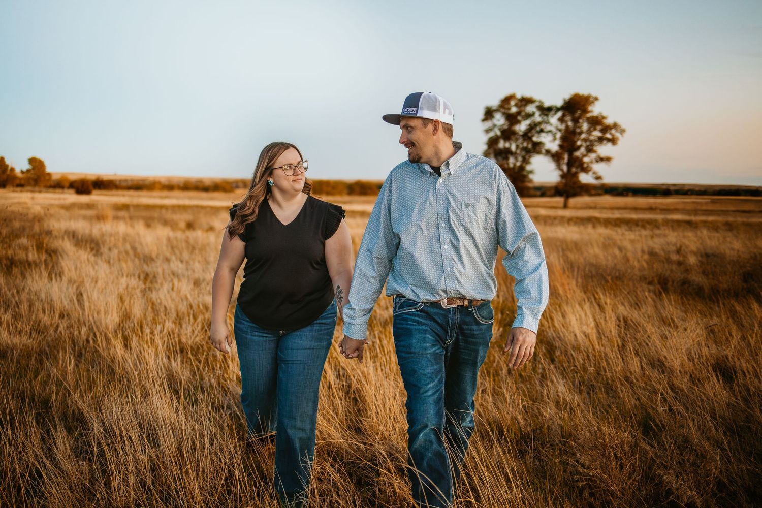 Couple holding hands walking through golden prairie field at sunset, wearing jeans and casual tops.