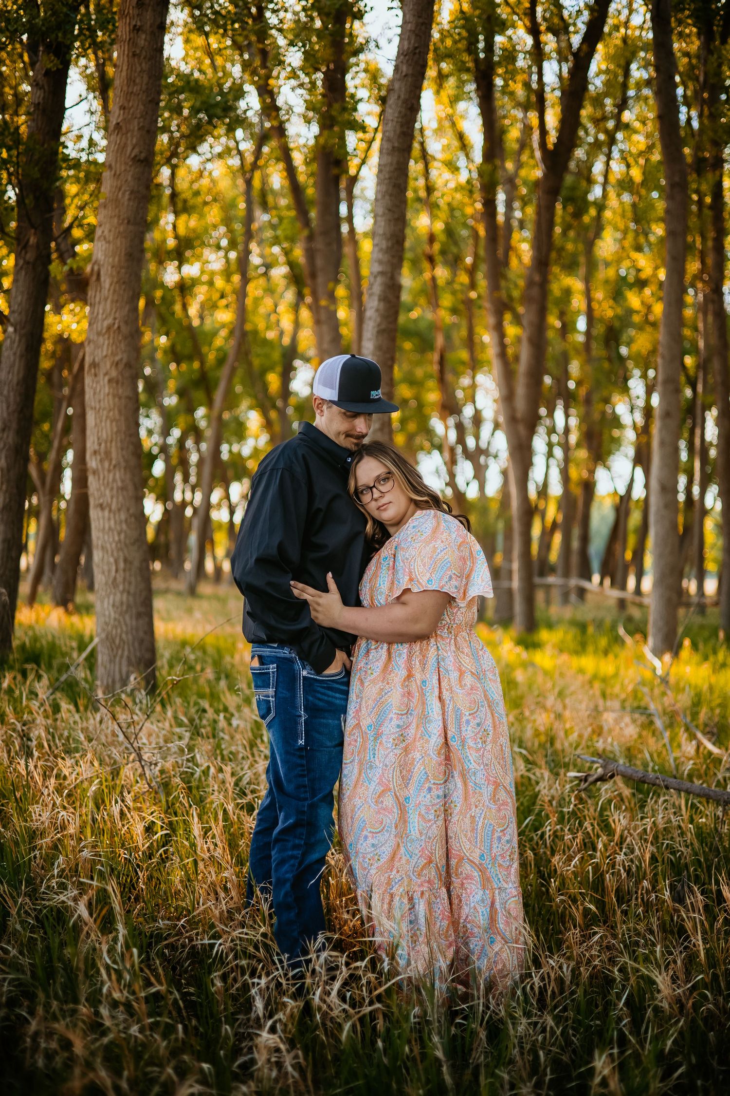 Couple embracing in golden sunlit forest, woman in floral dress and man in black hoodie and trucker hat, tall trees background.