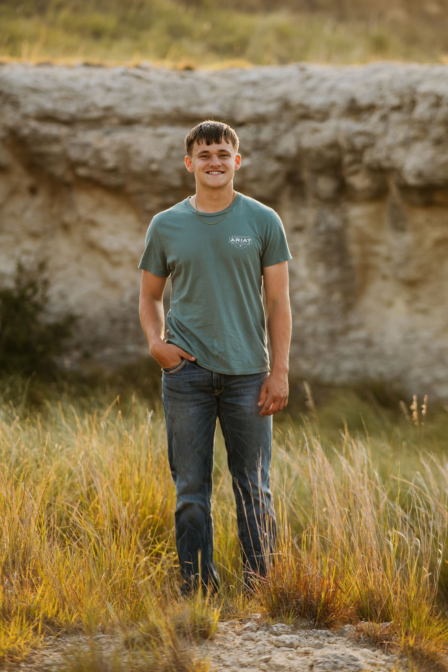 Person wearing a green t-shirt and jeans stands in a field with tall grass and rocky cliffs in the background.