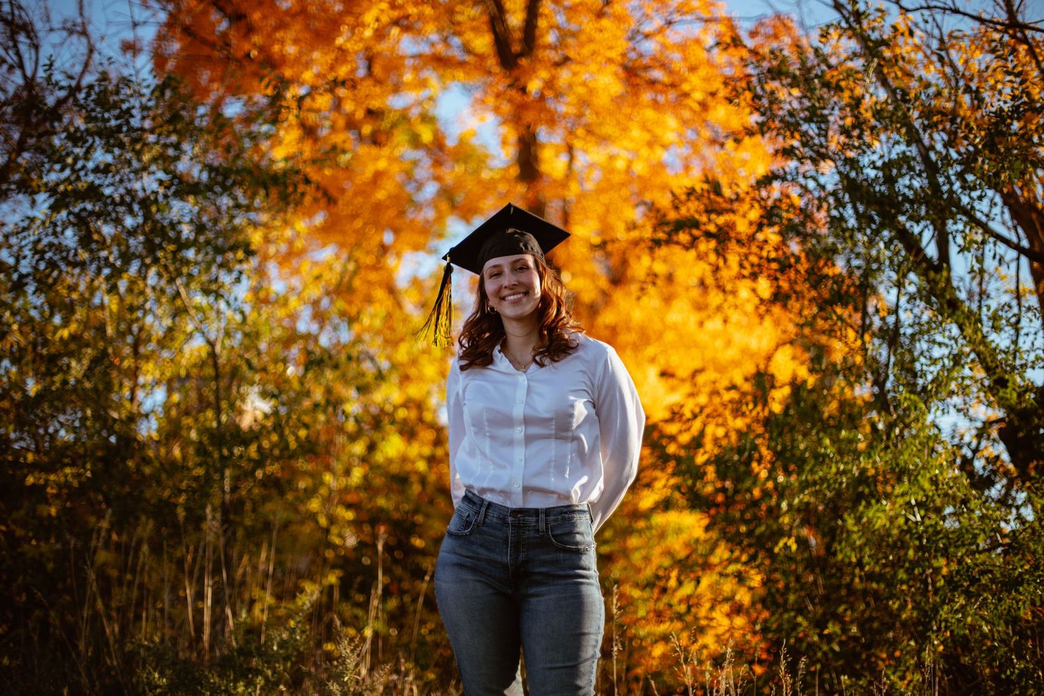 Graduate in cap and gown poses against vibrant fall foliage.