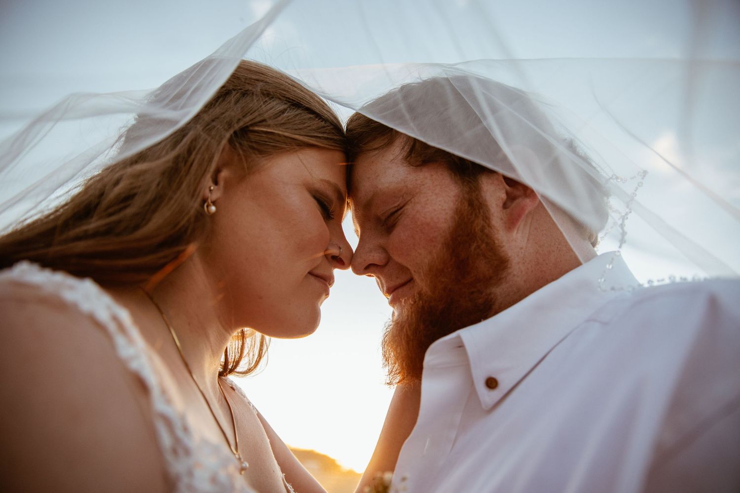 Romantic couple shares intimate moment under veil during sunset wedding photo session.