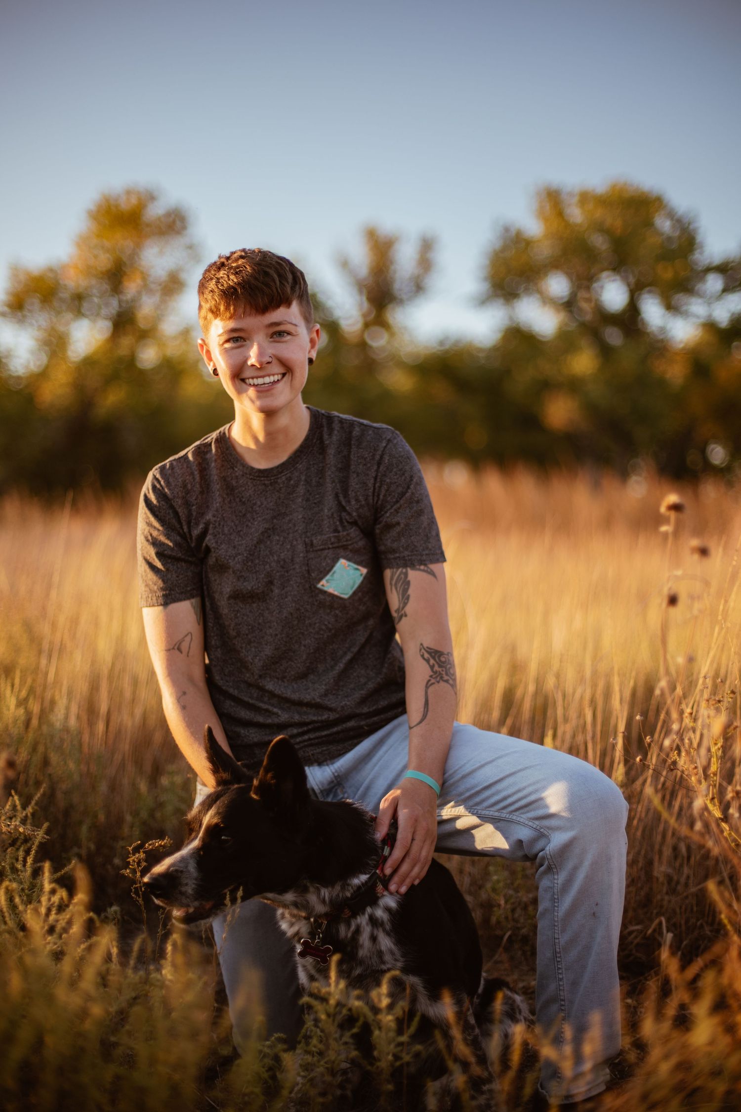 Person sitting with black dog in golden field during sunset wearing casual attire.