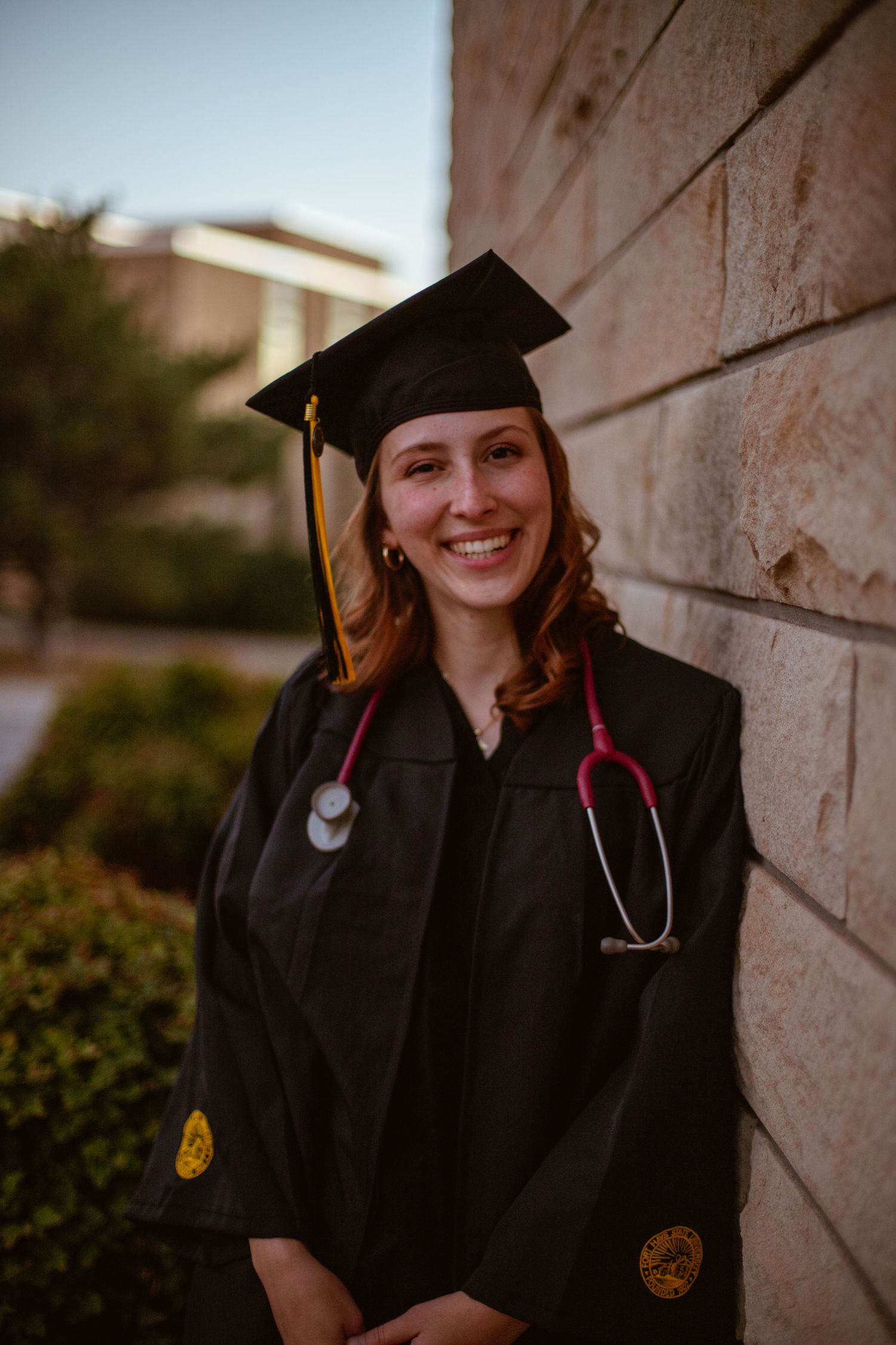 Medical graduate in cap and gown with stethoscope poses against stone wall.