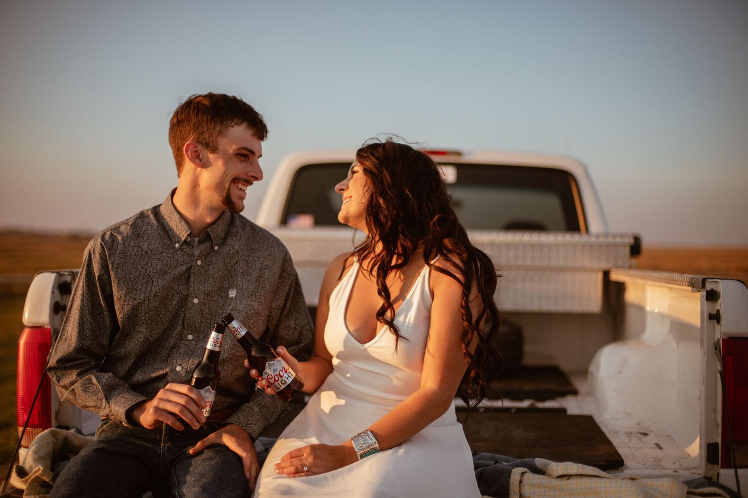 A couple shares drinks while sitting on the tailgate of a pickup truck at sunset.