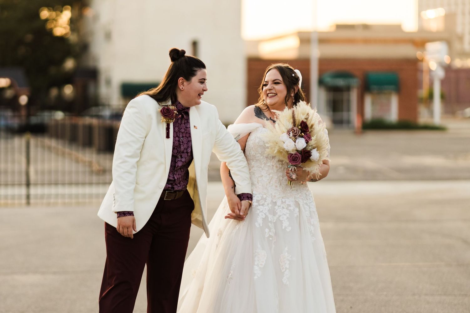 A couple in wedding attire shares a laugh while holding a floral bouquet.