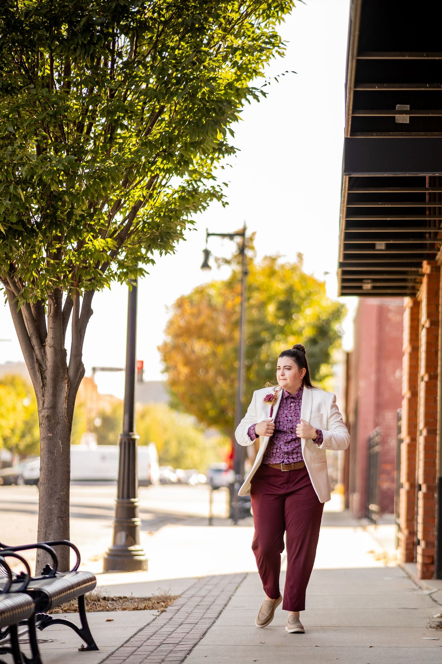 Person in white blazer and burgundy pants walking down urban sidewalk in autumn.