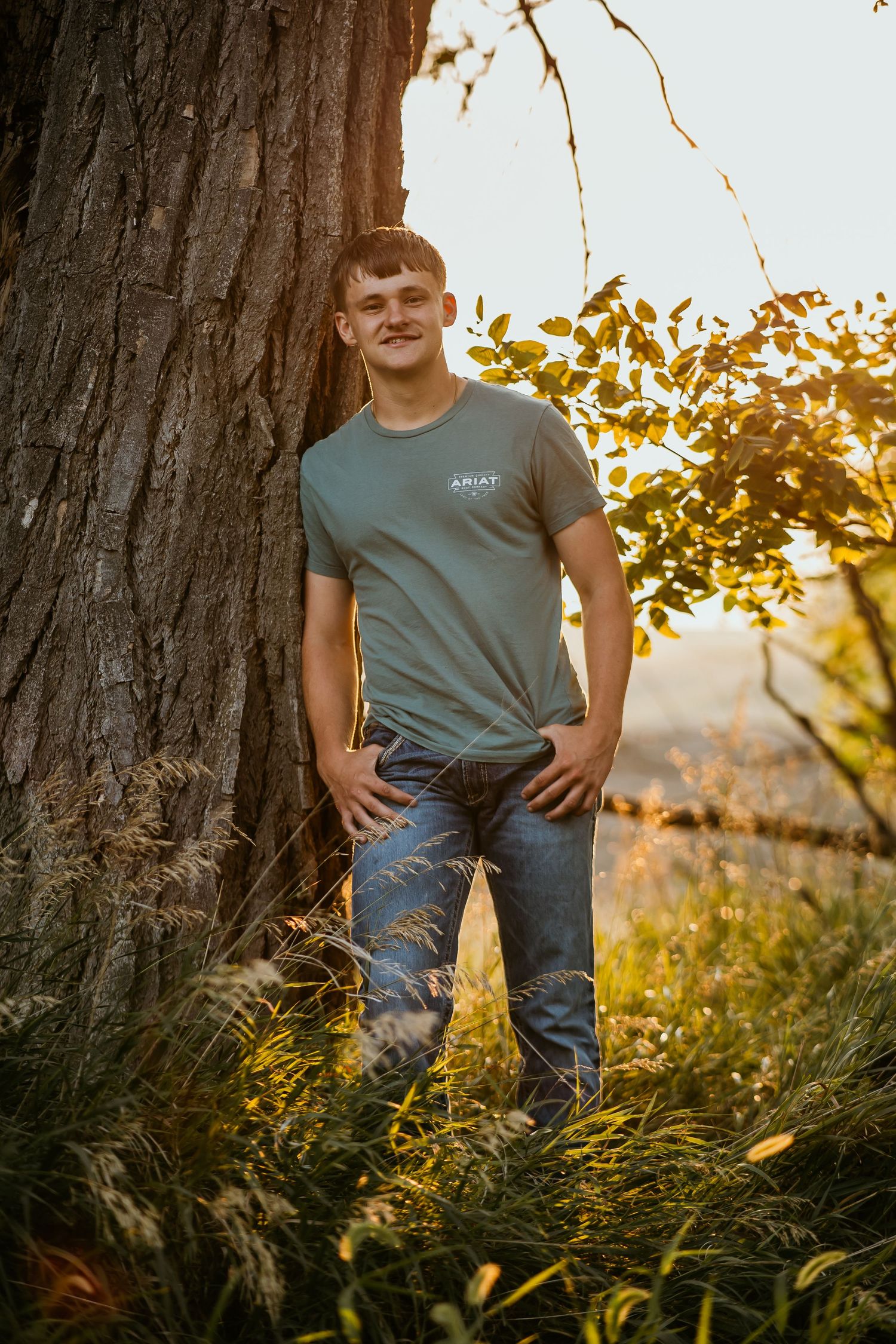 Person in gray t-shirt poses against large tree trunk during sunset in tall grass.