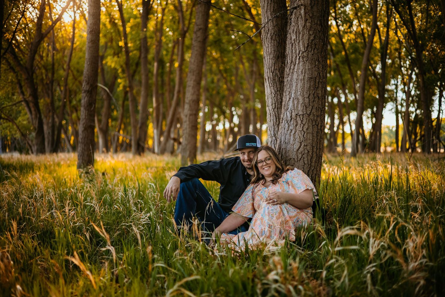 Couple sitting together against a large tree in a sunlit forest meadow with golden hour lighting.