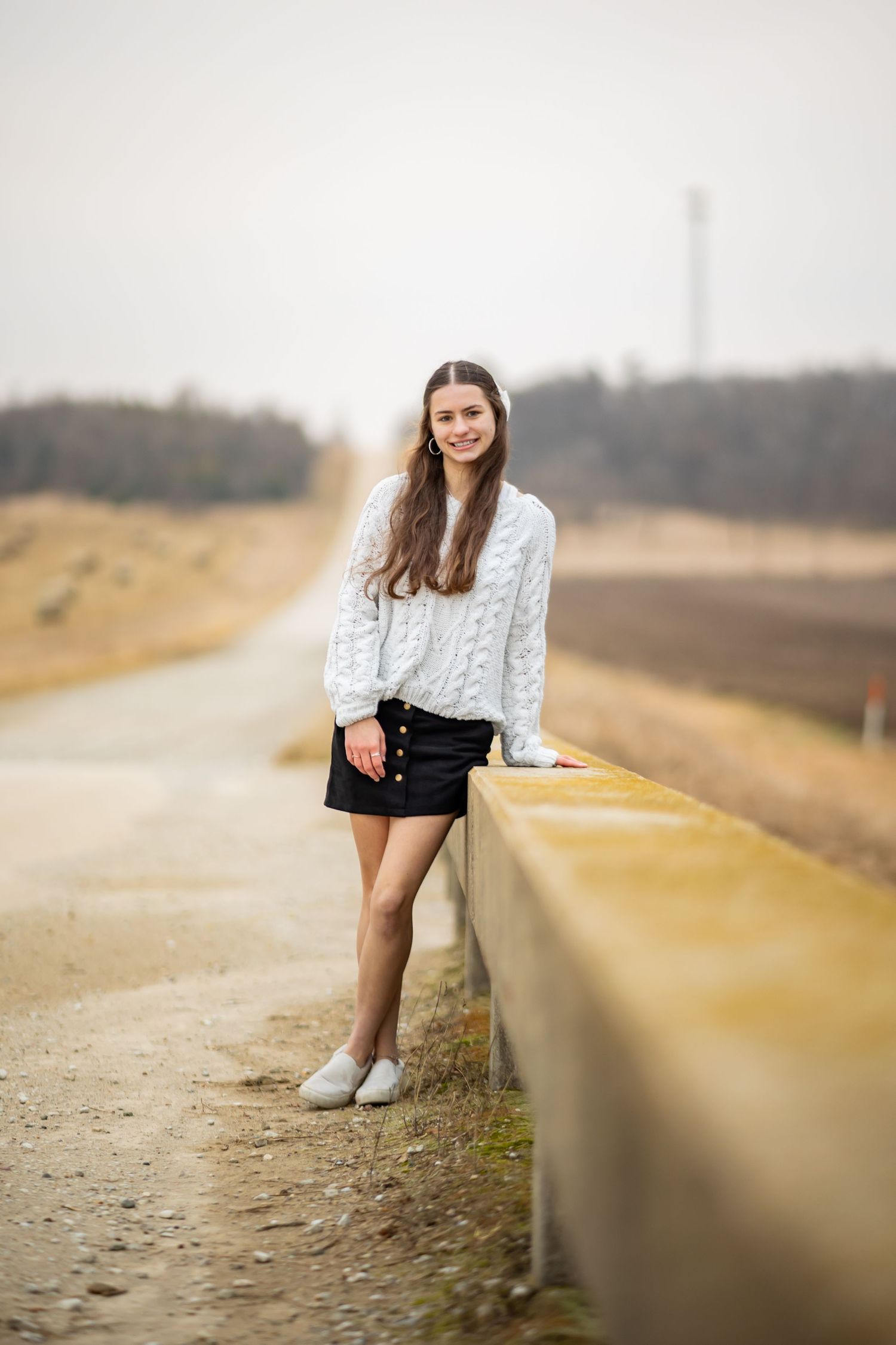 Young person in white sweater and shorts poses casually along a rural road.