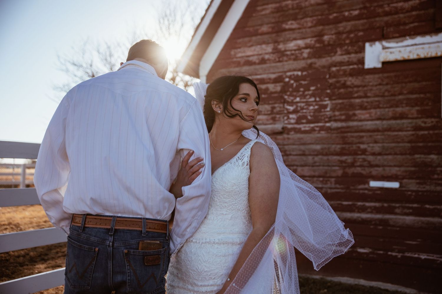 Romantic sunset wedding photo with couple embracing near rustic barn wall.