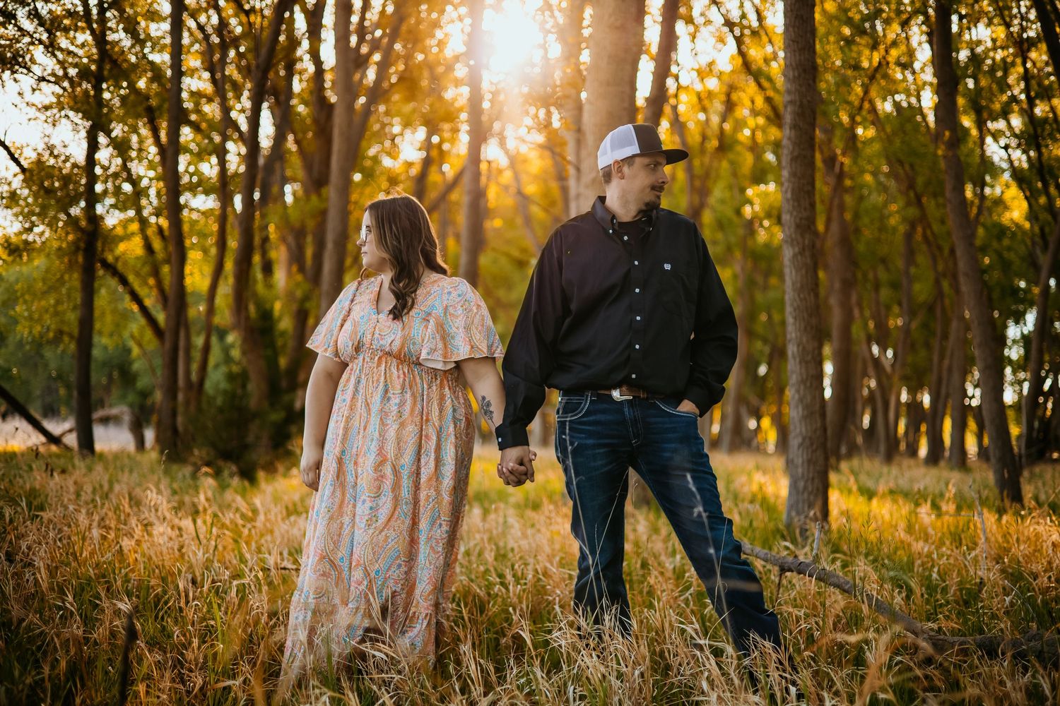 Couple holding hands in golden hour sunlight among tall grass and trees, woman in floral dress, man in black shirt and cowboy hat.