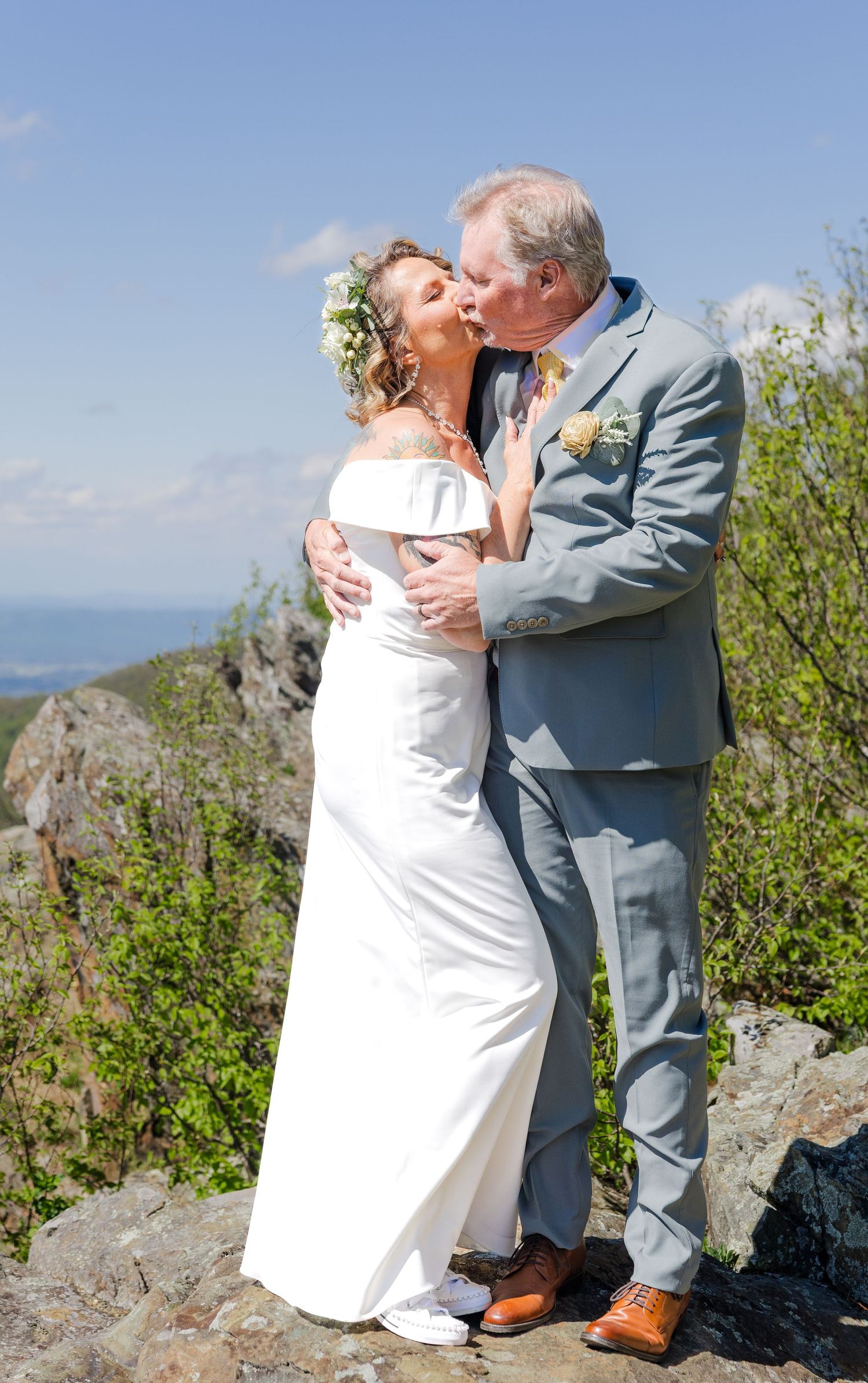 Bride and groom kissing on rocky mountain overlook, bride in white off-shoulder gown with floral crown, groom in light gray suit.