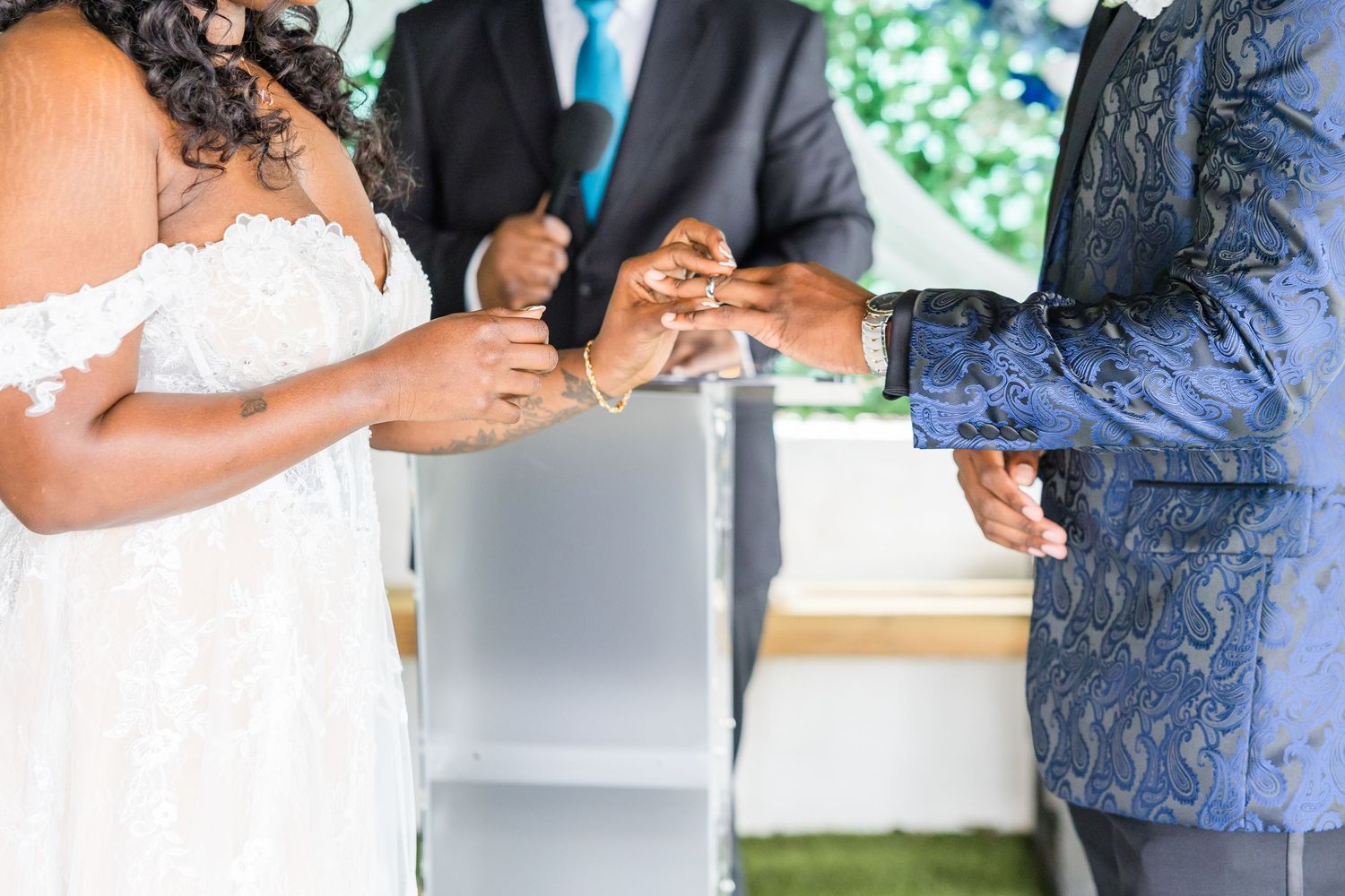 Close up of hands exchanging wedding rings during an outdoor ceremony with a white bridal gown visible.