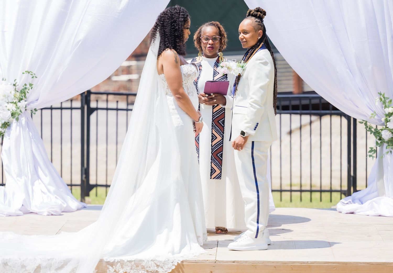 Outdoor wedding ceremony under white draped arch with couple and officiant at the altar.
