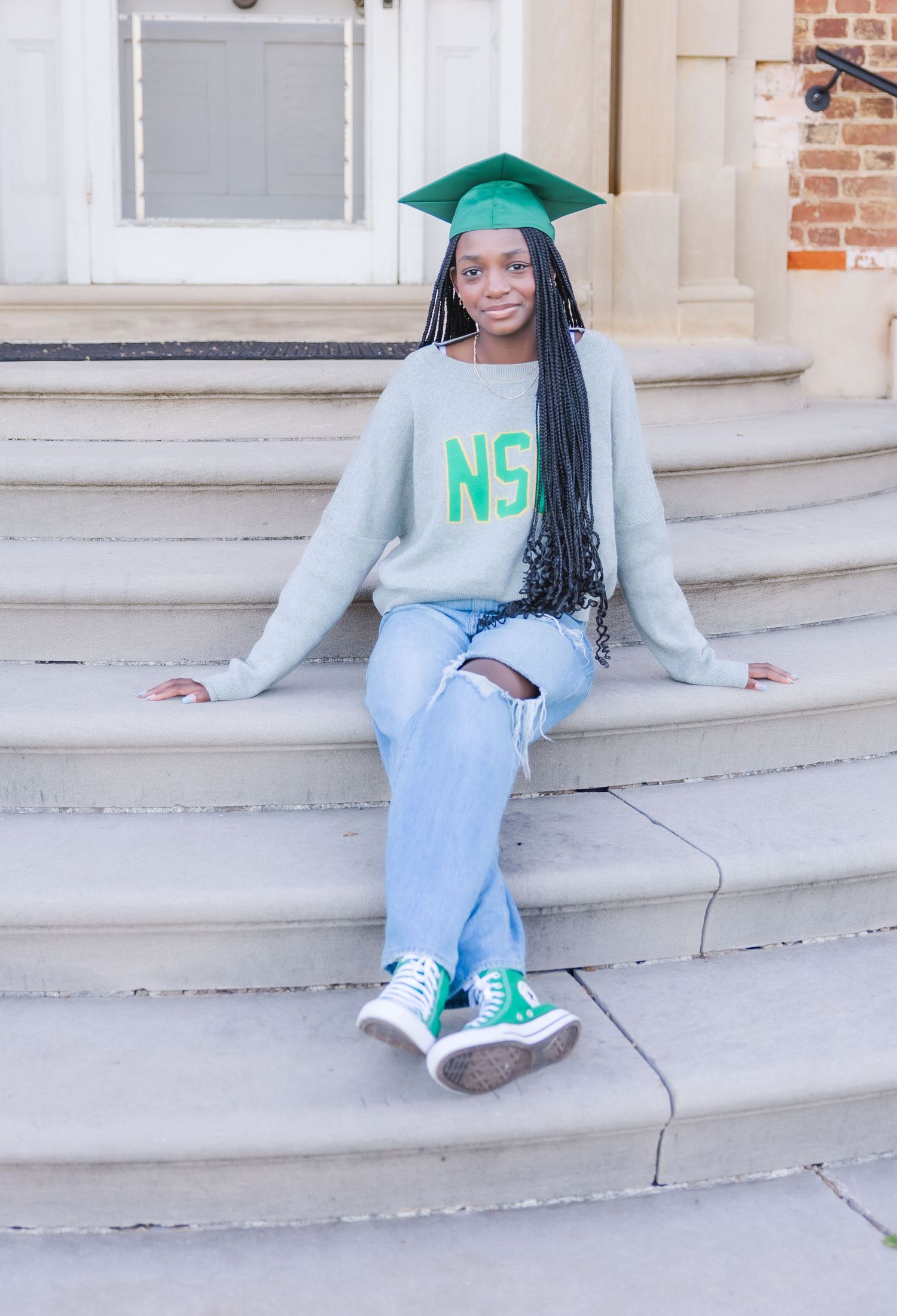 Graduate in green cap and NSU sweatshirt sits on white stone steps.