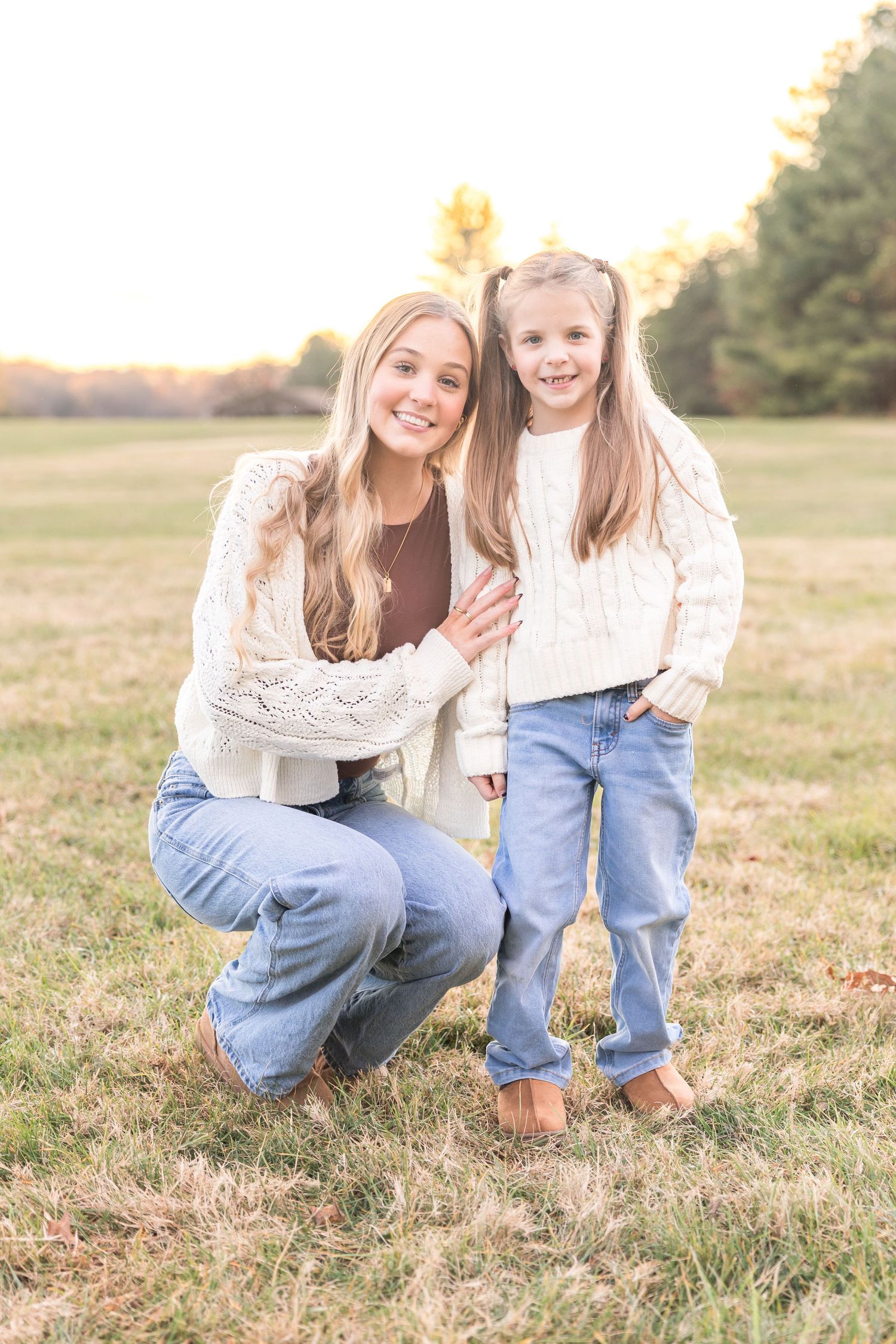 Two people in matching white tops and jeans sit together in a sunlit grassy field during golden hour.
