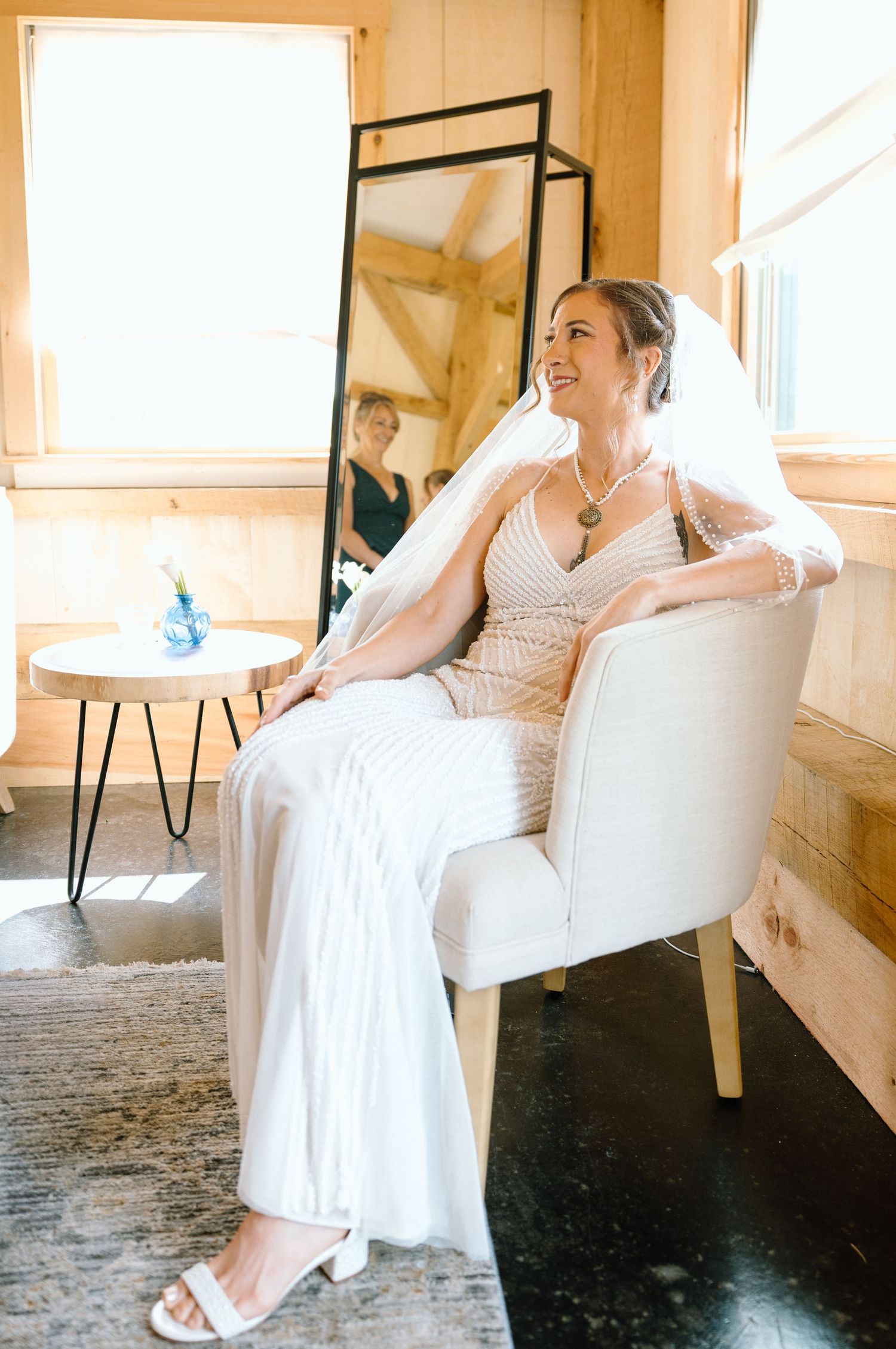 A smiling bride in a white beaded gown and veil sits in a cream chair in a sunlit rustic wooden room.