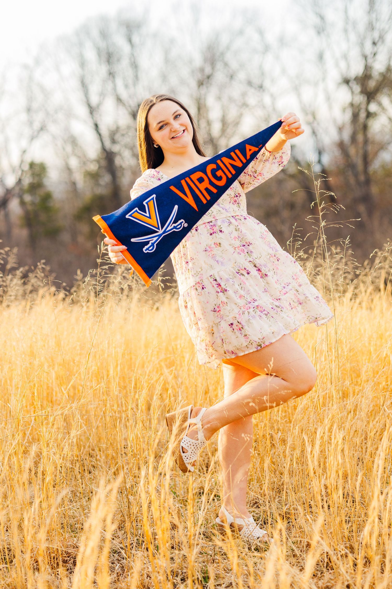 Student showing school spirit in a floral dress while holding a UVA pennant in a golden field at sunset.