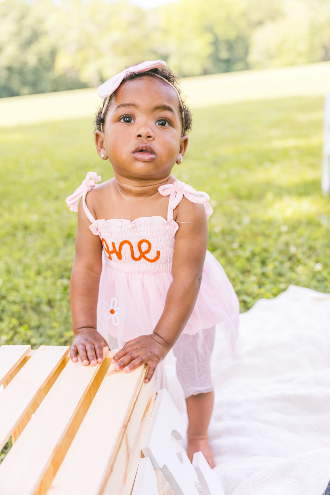 Baby wearing pink and orange dress sits on white blanket in grassy outdoor setting.
