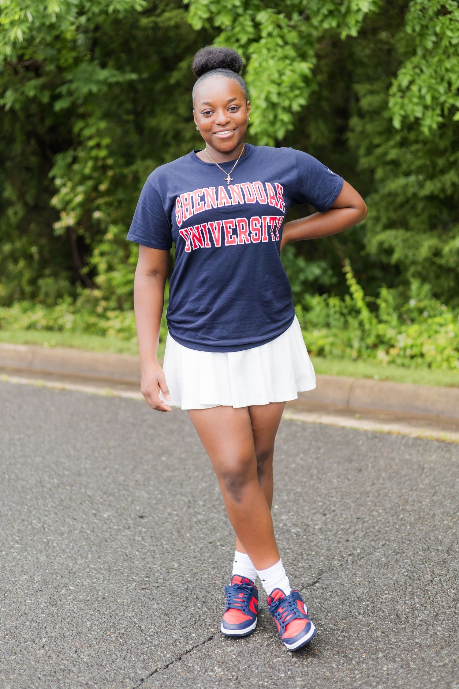 A person in university apparel strikes playful poses on pavement with trees in the background.