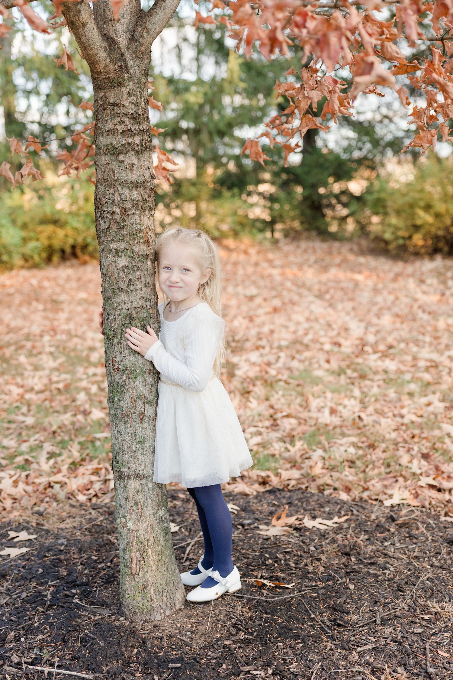 A child in a white sweater and navy pants plays near autumn trees with orange leaves and fallen foliage on the ground.