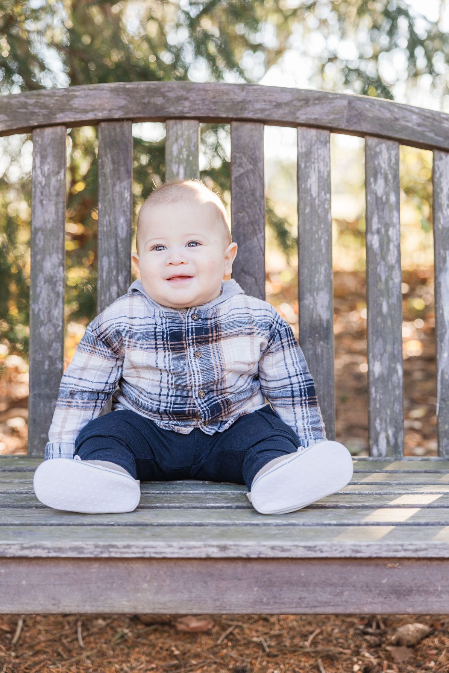A baby wearing a plaid shirt and white pants sits on a wooden bench in a park setting with trees in the background.